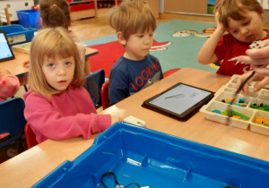 Dziewczynka i dwóch chłopców siedzi przy stoliku i próbują zbudować robocika z klocków Lego, a instrukcja wyświetla im się na tablecie.