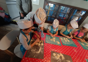 Dzieci siedzą przy stole i lepią pierogi z pomocą kucharza.