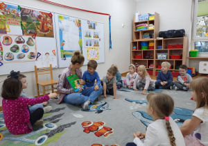 Dzieci siedzą na dywanie w kole, a mama Tomka prezentuje im książkę pt. "Tupcio Chrupcio. Umiem się dzielić"