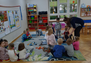 Dzieci siedzą na dywanie i czekają aż prowadząca zajęcia z robotyki wyjmie potrzebne do zajęć rzeczy.