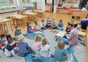 Dzieci siedzą na dywanie w kole i uważnie słuchają czytanego opowiadania.