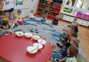 Dzieci siedzą na dywanie, rozmawiają z nauczycielką i poznają zmysły potrzebne do określenia cech owoców znajdujących się w miseczkach na stole.