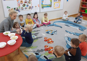Dzieci siedzą na dywanie, chłopiec z zasłoniętymi oczami za pomocą zmysłu węchu zgaduje co to za owoc.