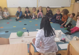 Dzieci siedzą w półkolu i obserwują jak prowadząca przedstawia im inny sposób na nadmuchanie balonika za pomocą ciepłej wody i drożdży.