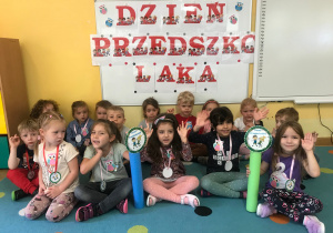 Dzieci siedzą na dywanie na tle napisu Dzień Przedszkola i prezentują medale Super Przedszkolaka.