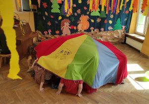 Dzieci siedzą na podłodze i chowają się pod chustą animacyjną.