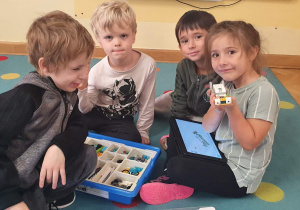 Trzech chłopców i dziewczynka siedzą na dywanie i prezentują robota wykonanego z klocków Lego na zajęciach z robotyki.
