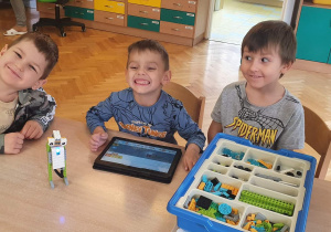 Trzech chłopców siedzi przy stole i prezentuje swojego robota wykonanego na zajęciach z robotyki z klocków Lego.