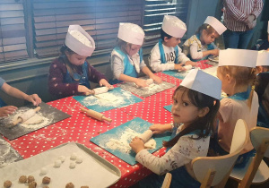 Dzieci siedzą przy stole ubrane w czapki i fartuchy kuchenne i wałkują ciasto na pierogi.