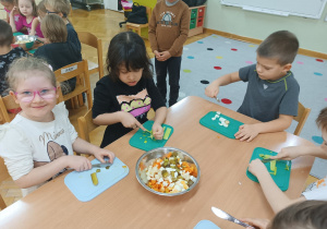 Dzieci siedzą przy stoliku, jedna dziewczynka kroi pora, druga ogórka, a chłopiec kroi jajko.