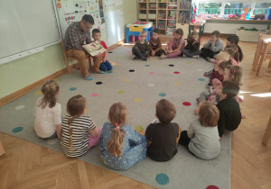 Dzieci siedzą w półkolu na dywanie przed nimi tata Jasia czyta im bajkę pt."Pucio w mieście".