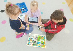 Dwie dziewczynki i chłopiec siedzą na dywanie i próbują zbudować robota z klocków Lego na zajęciach z robotyki.
