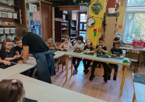 Dzieci siedzą przy stołach, a prowadząca pokazuje im jak mają lepić z gliny.