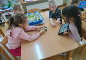 Dzieci siedzą przy stoliku i za pomocą instrukcji wyświetlanej na tablecie starają się zbudować robota z klocków Lego na zajęciach z robotyki.