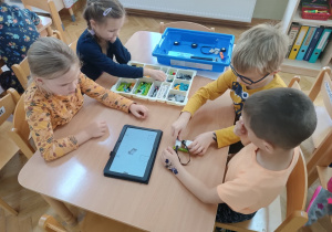 Dzieci siedzą przy stoliku i za pomocą instrukcji wyświetlanej na tablecie starają się zbudować robota z klocków Lego na zajęciach z robotyki.