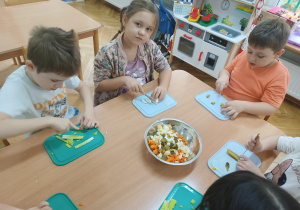 Dzieci siedzą przy stoliku, chłopcy kroją pora i ogórki, a dziewczynka kroi jajko do sałatki.