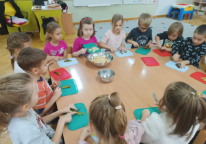 Dzieci siedzą przy stoliku i kroją ogórka , a jedna dziewczynka dodaje wszystkie warzywa do sałatki jarzynowej.