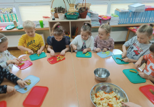 Dzieci siedzą przy stoliku i kroją ogórka do sałatki jarzynowej.