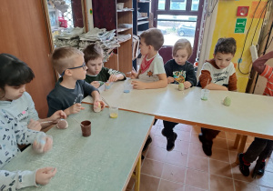 Dzieci siedzą przy stolikach i dekorują figurki z gliny.