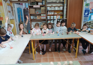 Dzieci siedzą przy stolikach i w skupieniu malują ulepione z gliny figurki jeżyków.