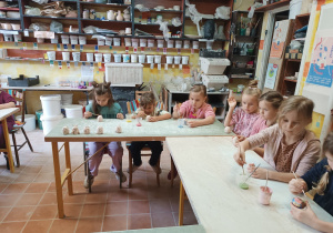 Dzieci siedzą przy stolikach i w skupieniu malują wcześniej ulepione z gliny figurki jeżyków.