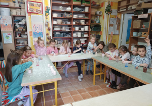 Dzieci siedzą przy stolikach i w skupieniu malują wcześniej ulepione z gliny figurki jeżyków, a kilkoro dzieci podnosi rączki do góry.