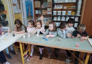 Dzieci siedzą przy stolikach i w skupieniu malują wcześniej ulepione z gliny figurki jeżyków, a jedna dziewczynka podnosi rączkę do góry.