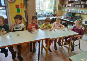 Dzieci siedzą przy stolikach i w skupieniu malują wcześniej ulepione z gliny figurki jeżyków.