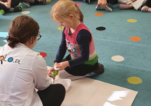 Dziewczynka siedzi na dywanie I pomaga Pani instruktorce zrobić doświadczenie z kolorowymi kuleczkami.
