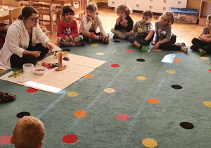 Pani prowadząca warsztaty Labolo pokazuje dzieciom siedzącym w kole na dywanie przedmioty, które przyniosła jak np.: koronę, kulki, wagę.