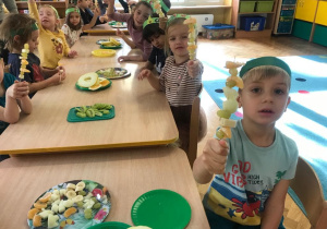 Dzieci siedzą przy stolikach i prezentują swoje owocowe szaszłyki.
