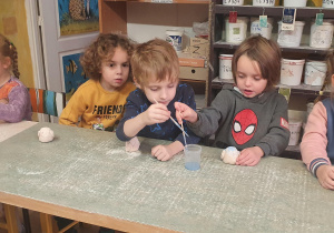 Dzieci siedzą przy stole w pracowni ceramicznej i malują farbkami sówki z gliny.