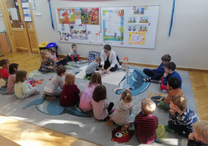 Dzieci siedzą na dywanie w półkolu, a jeden z chłopców wspólnie z prowadzącą przesypuje plastikowe koraliki do pojemniczka.