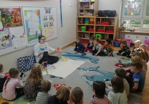 Dzieci siedzą na dywanie i słuchają opowiadania o Archimedesie, a prowadząca pokazuje im obrazek ze słynnymi słowami Archimedesa: "Eureka".