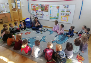 Dzieci siedzą w kole na dywanie z prowadzącą warsztaty z robotyki wita się z dziećmi.