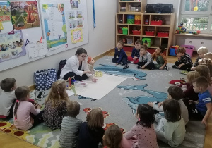 Dzieci siedzą na dywanie w półkolu, a prowadząca warsztaty pokazuje im dwie kostki.