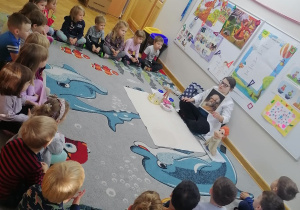 Dzieci siedzą w półkolu na dywanie, a prowadząca warsztaty pokazuje im portret Archimedesa.