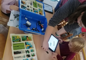 Dzieci siedzą przy stoliku i z pomocą prowadzącej warsztaty próbują zbudować robota z klocków Lego na zajęciach z robotyki.