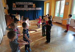 Dzieci stoją w rozsypce na sali gimnastycznej z rękami wyciągniętymi przed siebie i "jadą konno".