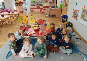 Dzieci siedzą w kółku dookoła stolika z miską z owocami i trzymają ramkę do fotobudki.