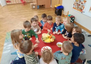 Dzieci siedzą w kółku dookoła stolika, na którym znajduje się miska pełna owoców i próbują je nazwać.