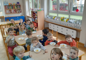 Dzieci siedzą przy stolikach i zajadają się samodzielnie wykonaną sałatką owocową i unoszą kciuki w górę.