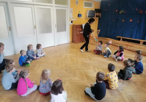 Dzieci siedzą w kole na zajęciach z rytmiki, a Pani pokazuje im bębenek i podaje jednemu z chłopców, który ma powtórzyć rytm za nią.