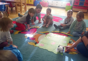 Dzieci siedzą na dywanie w półkolu i obserwują jak prowadząca zajęcia z robotyki i programowania ustawia i programuje robocika pandę.