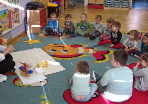 Dzieci siedzą na dywanie w półkolu, a prowadząca warsztaty chemiczne LABOLO pokazuje im pojemniczki z wodą i dzieci próbują zgadnąć, w którym pojemniczku jest więcej cieczy.