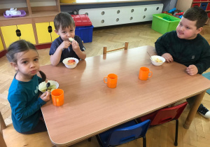 Chłopcy i dziewczynka siedzą przy stoliku i jedzą owoce z miseczek na drugie śniadanie.