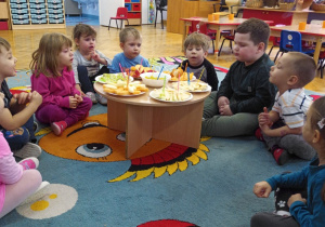 Dzieci siedzą w kółku przy stoliku i oglądają owoce przygotowane z okazji Światowego Dnia Owoców i Warzyw.
