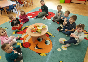 Dzieci siedzą w kole, na środku stoi stolik ze smacznymi owocami z okazji Światowego Dnia Warzyw i Owoców.
