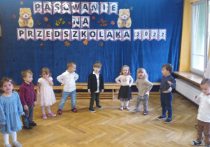Dzieci stoją w półkolu i śpiewają i tańczą do przygotowanej piosenki.