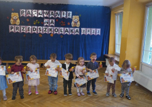 Dzieci stoją ustawione w rzędzie w rękach trzymając dyplomy i prezenty, które otrzymały podczas pasowania na przedszkolaka.